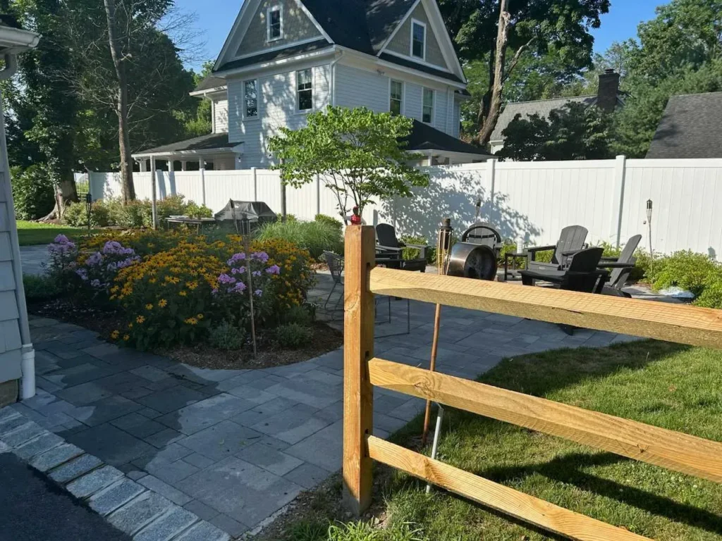 Garden area with flowers, patio furniture, and a white fence beside a house.